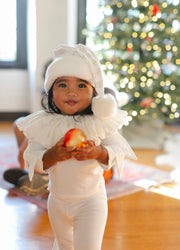 Child in a white outfit holding an apple with a Christmas tree in the background