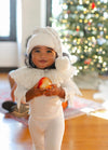 Child in a white outfit holding an apple with a Christmas tree in the background